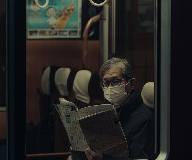 A senior man wearing a mask reads a newspaper on a Tokyo train, Japan, capturing a moment of daily life.